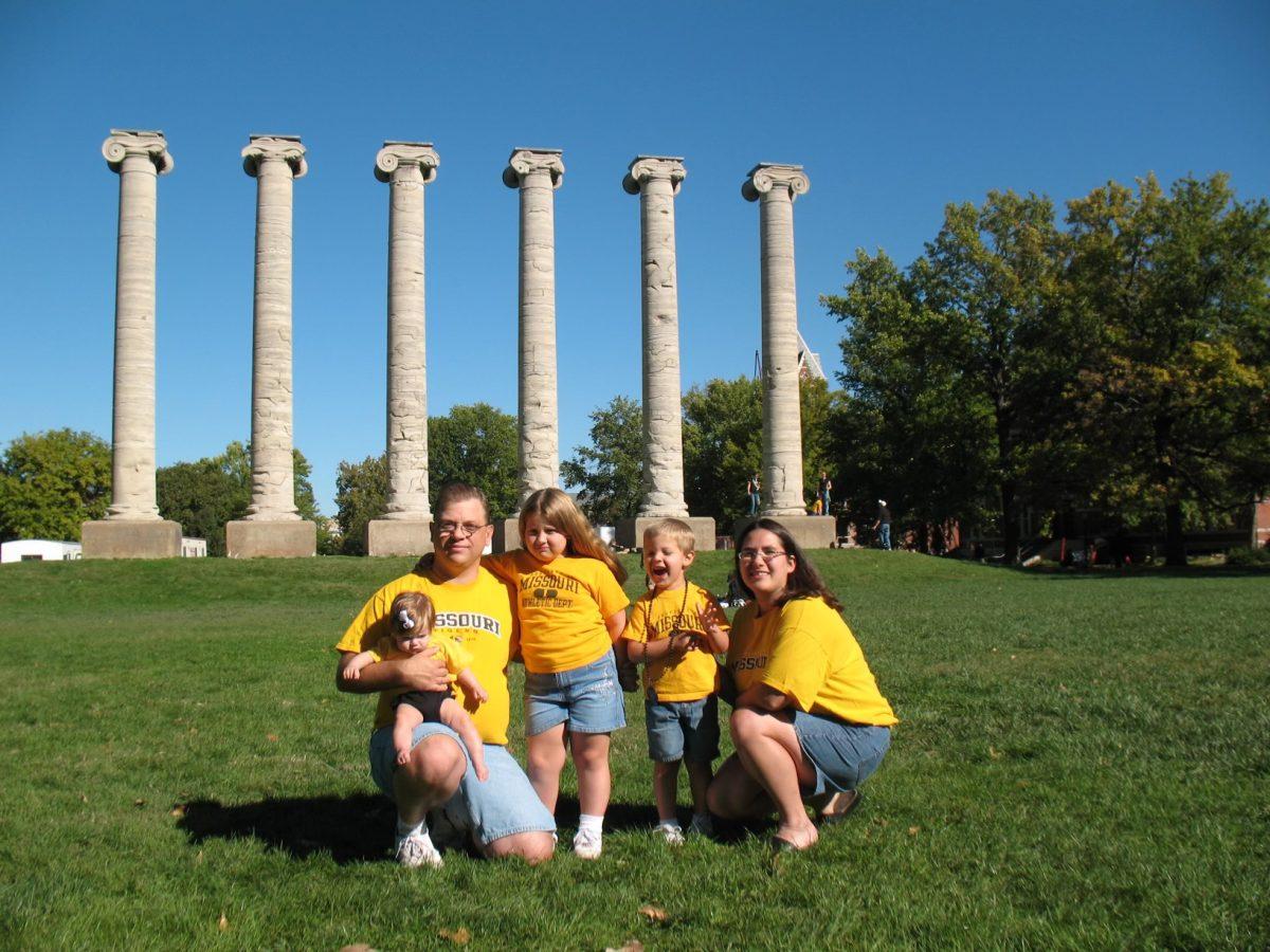 A family of five poses in front of the columns wearing jeans and gold Mizzou themed shirts.