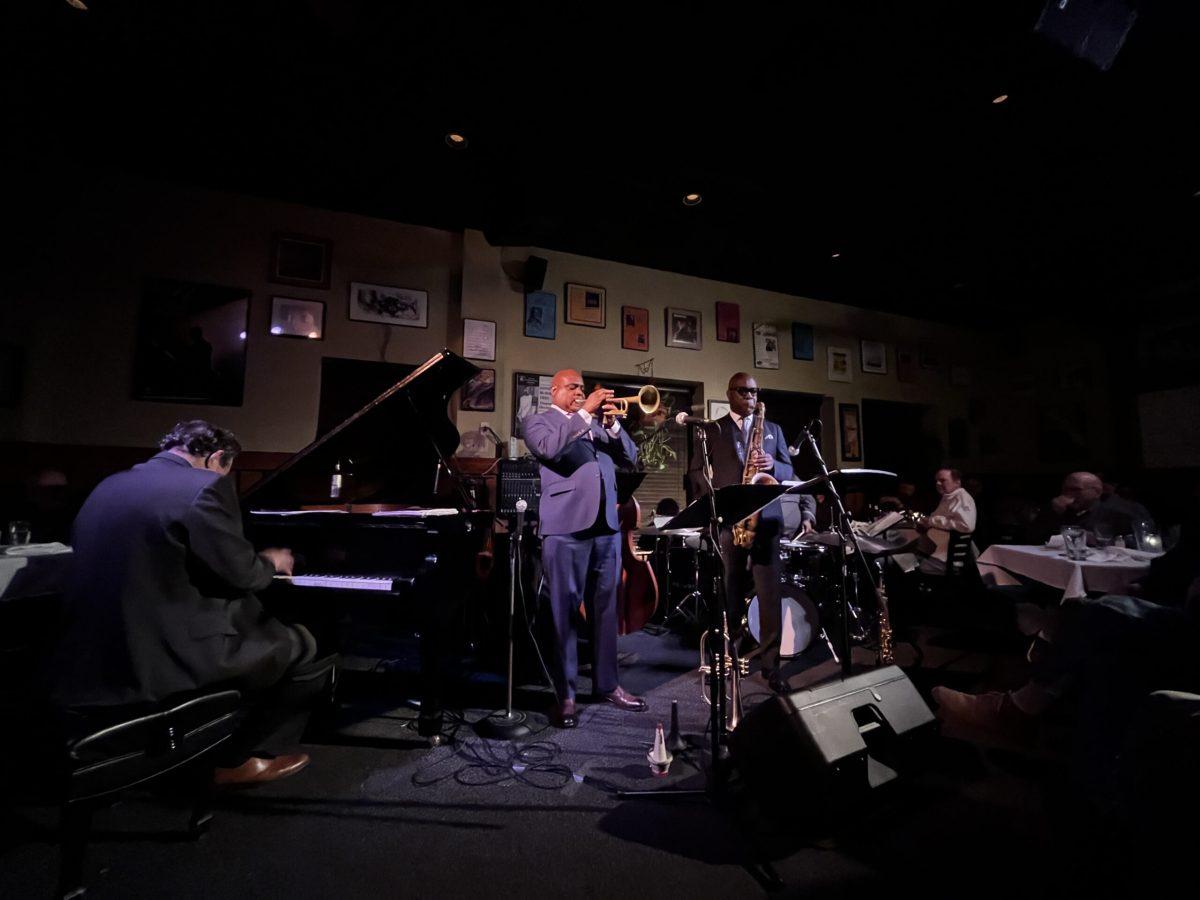 Terell Stafford plays trumpet on a cramped, dark stage with other band members playing piano and saxaphone.