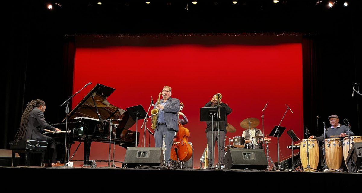 Elio Villafranca plays saxaphone with his band on a stage with a red-lit background.