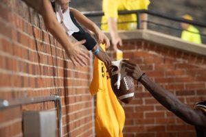 The hands of football game attendees reach over a stone wall holding items for a football player to sign.