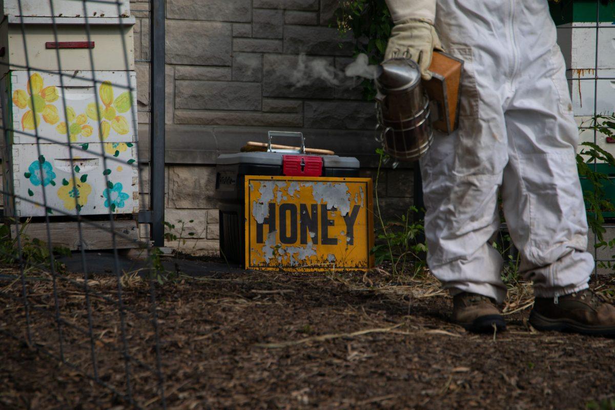 A weathered, yellow sign that reads “HONEY” in black ink leans against a tool box. On the right, George Frees stands facing away from the sign with a smoker in his right hand.