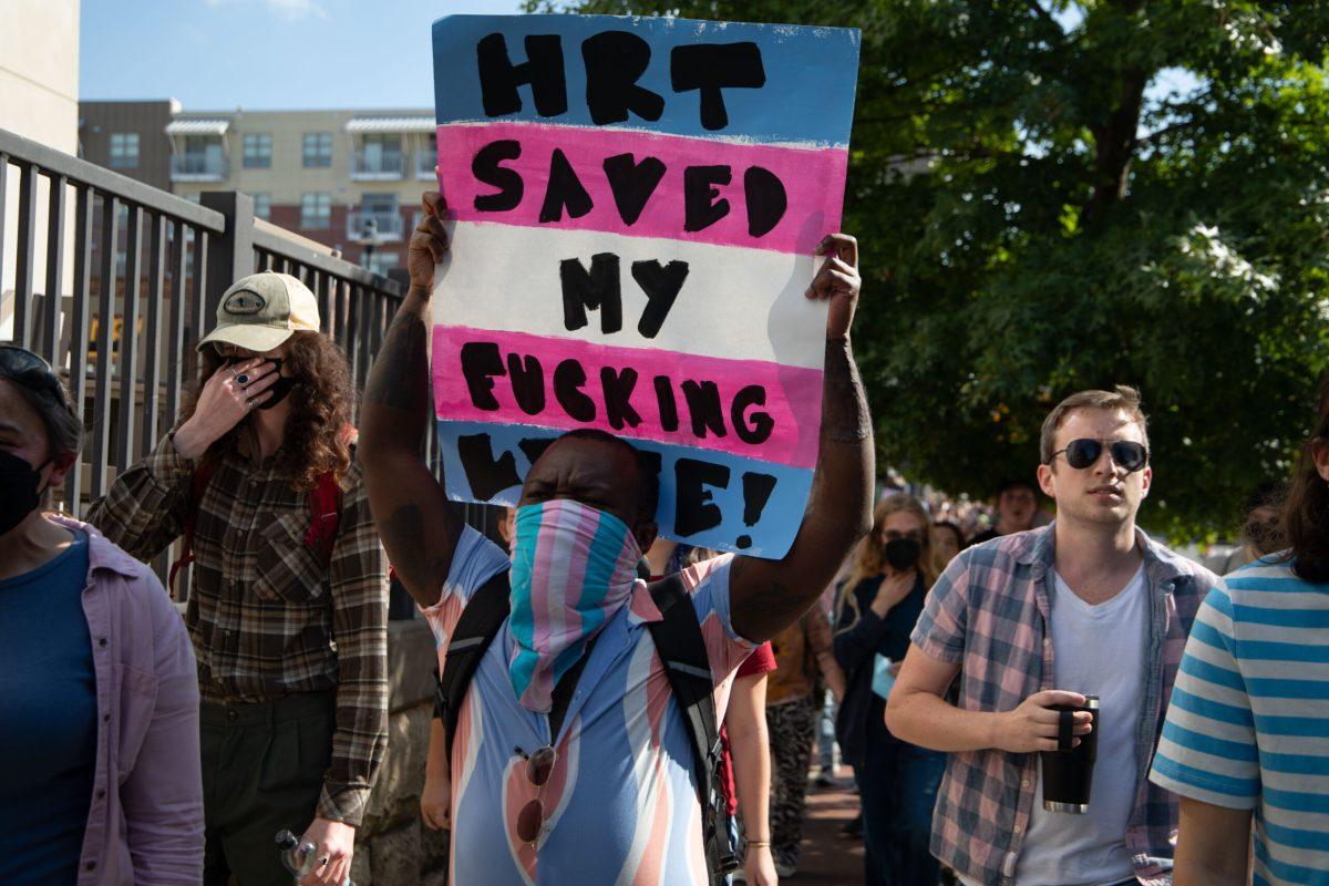 Kofi Opam holds up a blue, pink and white protest sign which reads “HRT SAVED MY FUCKING LIFE!” written in black paint. 