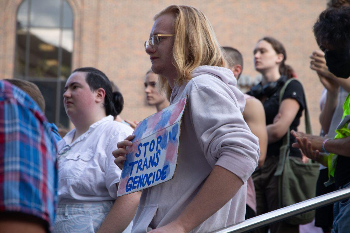 Kyle Maki holds a pink, blue and white protest sign that says “STOP TRANS GENOCIDE” in blue paint.
