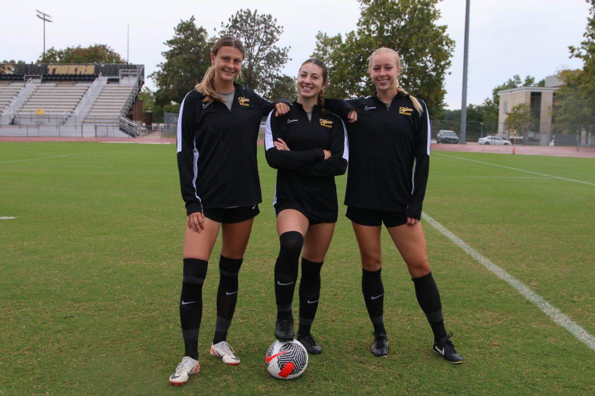 Three soccer players posing for a photo while the middle player holds a soccer ball.