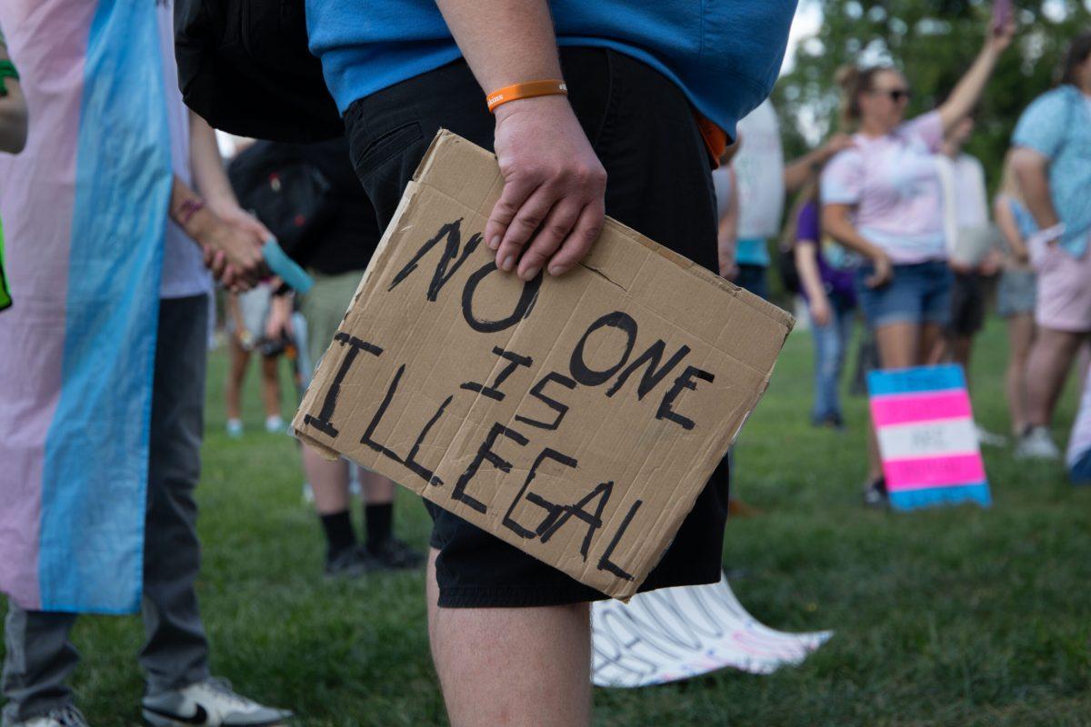 A protester holds a small cardboard sign at their side with the words “NO ONE IS ILLEGAL” written in black marker.