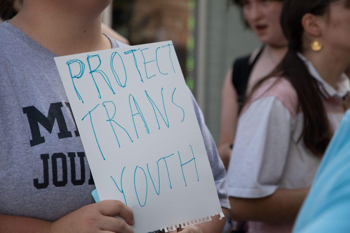 A protester holds up a white sign to their chest that reads “PROTECT TRANS YOUTH” in light blue ink.