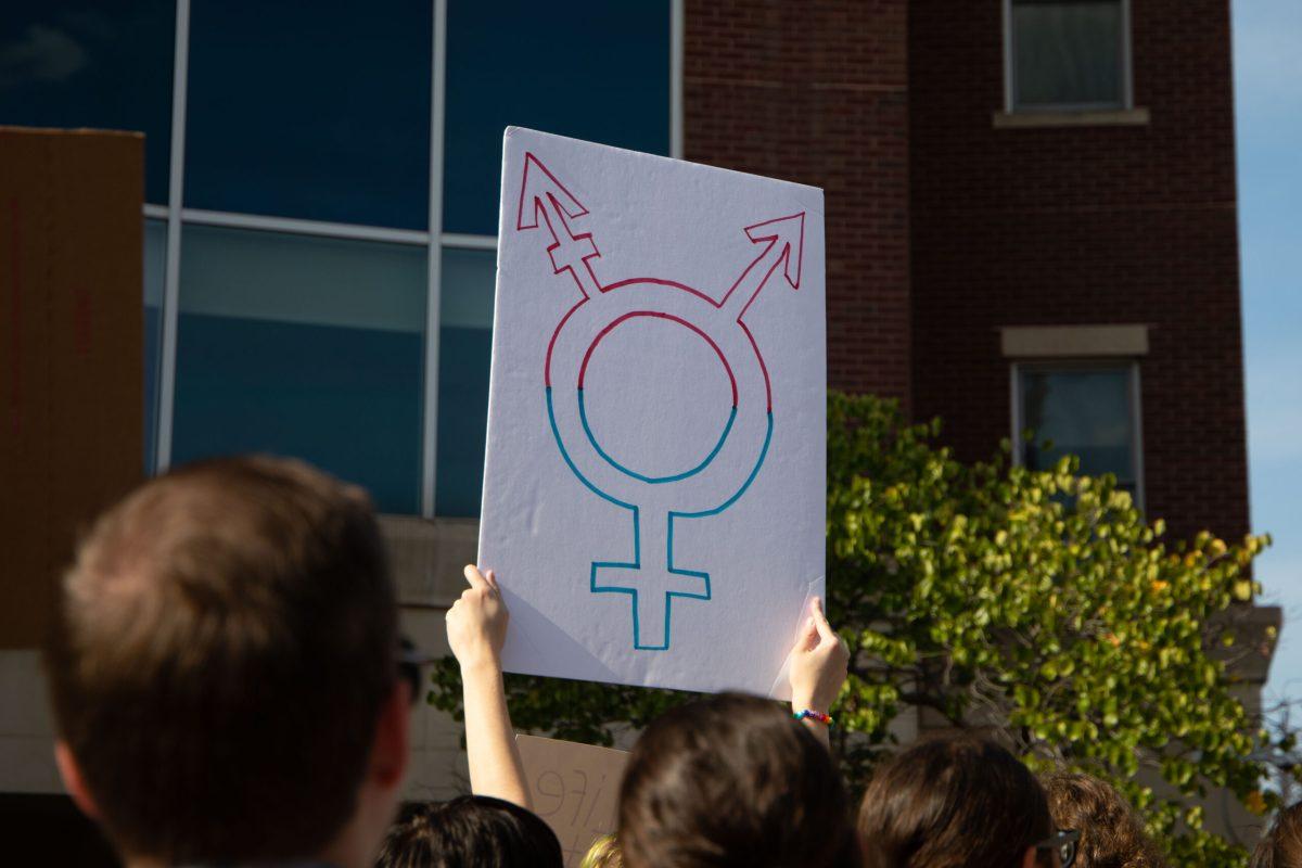 A protestor holds up a white sign with the trans symbol written on it in pink and blue ink in front of Columbia City Hall.