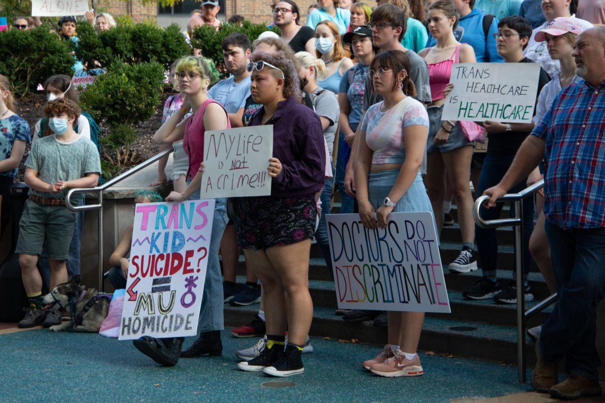 Protesters stand on the steps of Columbia City Hall while holding protest signs. A sign on the left reads “TRANS KID SUICIDE? MU HOMICIDE.” A sign in the middle reads “My life is NOT a crime.” A sign on the right reads “DOCTORS DO NOT DISCRIMINATE.”