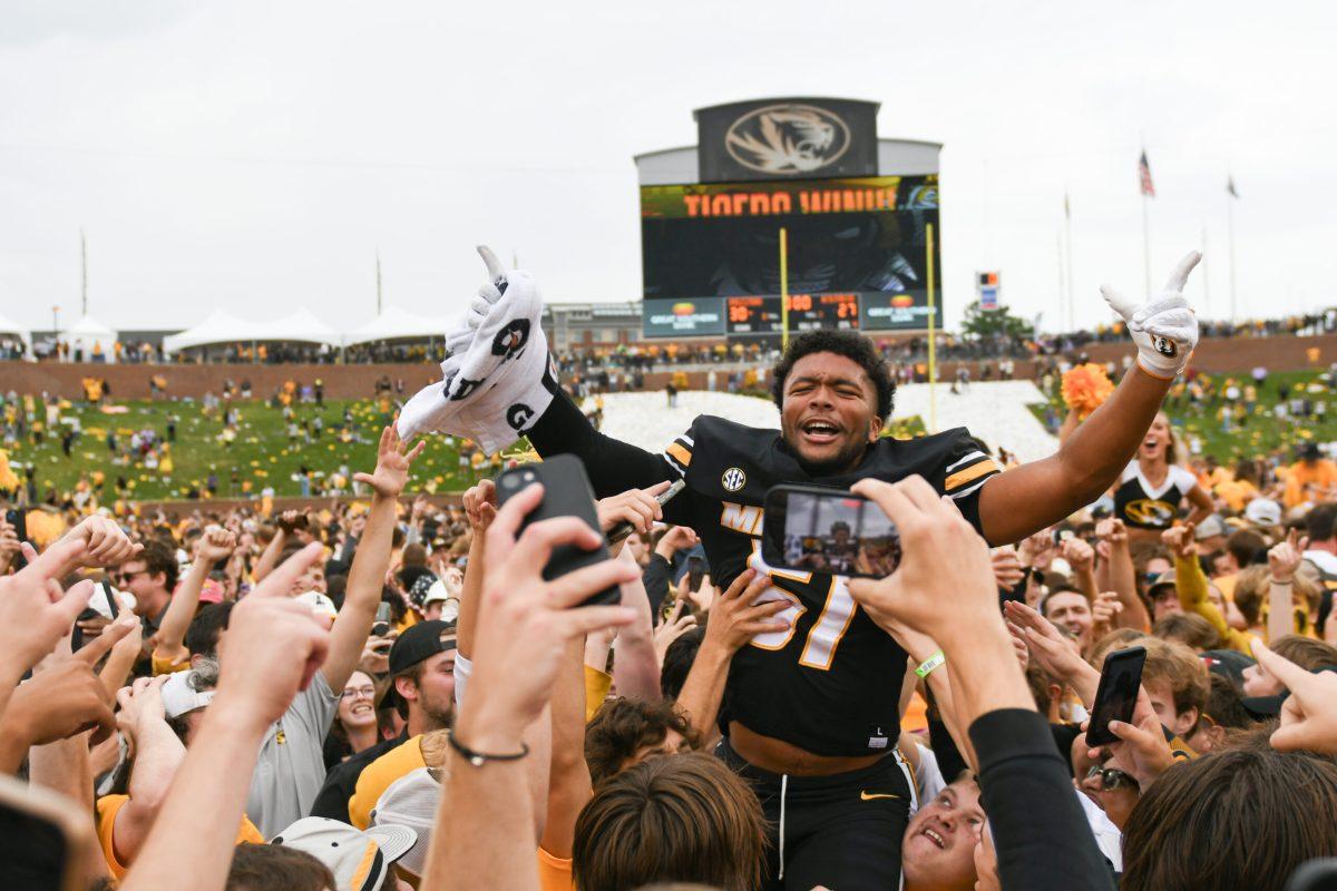 A large crowd of Missouri fans hold up a football player, who points his fingers at the crowd.