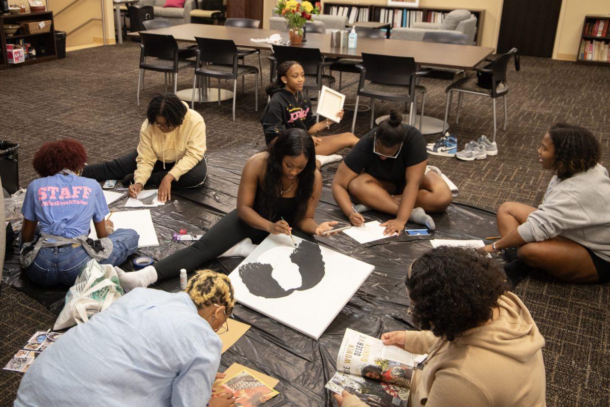 Eight students sit on a plastic floor protector while working on various artistic projects including collaging, painting and drawing. Central to the image, a guest at the event paints on a large canvas.