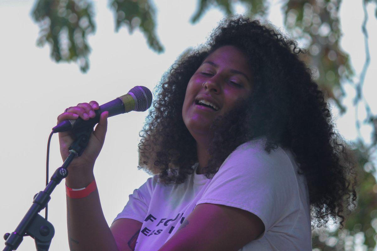 A singer holds a microphone while singing with head slightly tilted facing the camera.