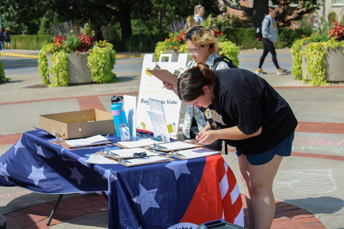 Two students writing on a clipboard registering to vote next to a booth decorated for Voter Registration Day.