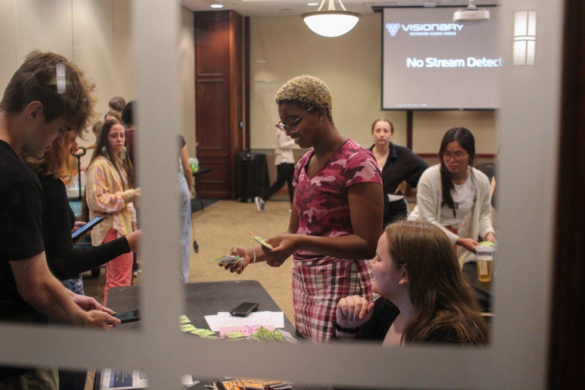 Event coordinators pass name tags to students as they stand in line in front of a table. The photo is taken through a window.