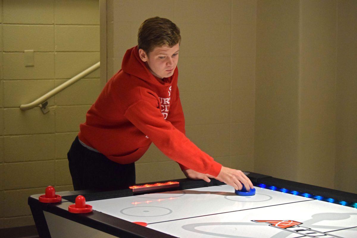 A student is playing air hockey with a blue mallet. Their arm is extended over the table in the opposite direction of where the puck is located.