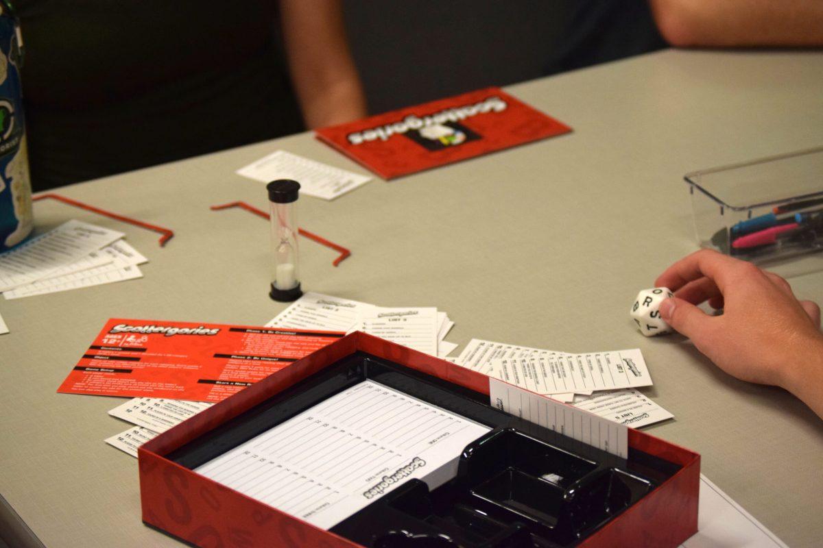 An open box of “Scattergories” is seen on a table with category cards splayed around the box. A hand is reaching for a 20-sided die with a different letter displayed on each side.