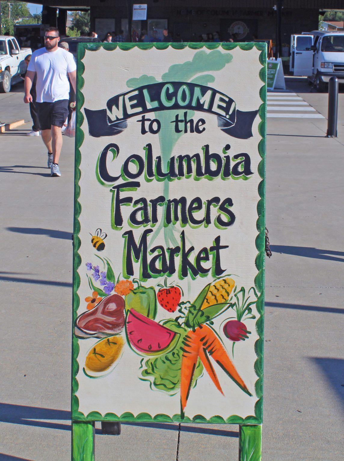 A “Welcome to the Columbia Farmers Market” sign stands in front of the farmers market.