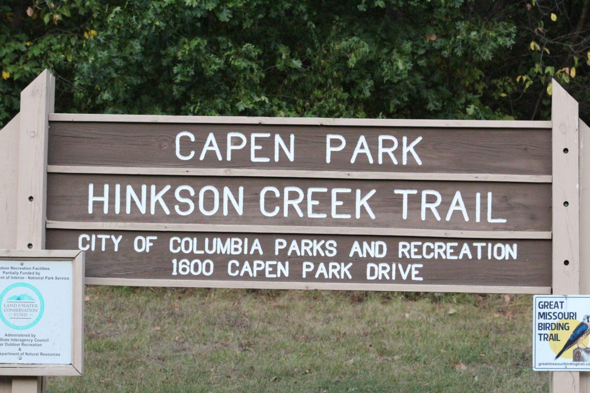 A sign reading “Capen Park Hinson Creek Trail” sits in front of trees.
