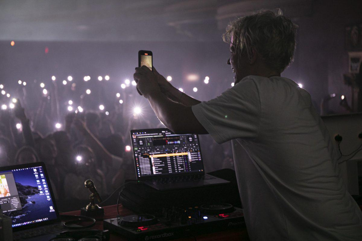 A man, facing the crowd, holds a phone with his hands outstretched while the crowd holds phones with their flashlights on above their heads.