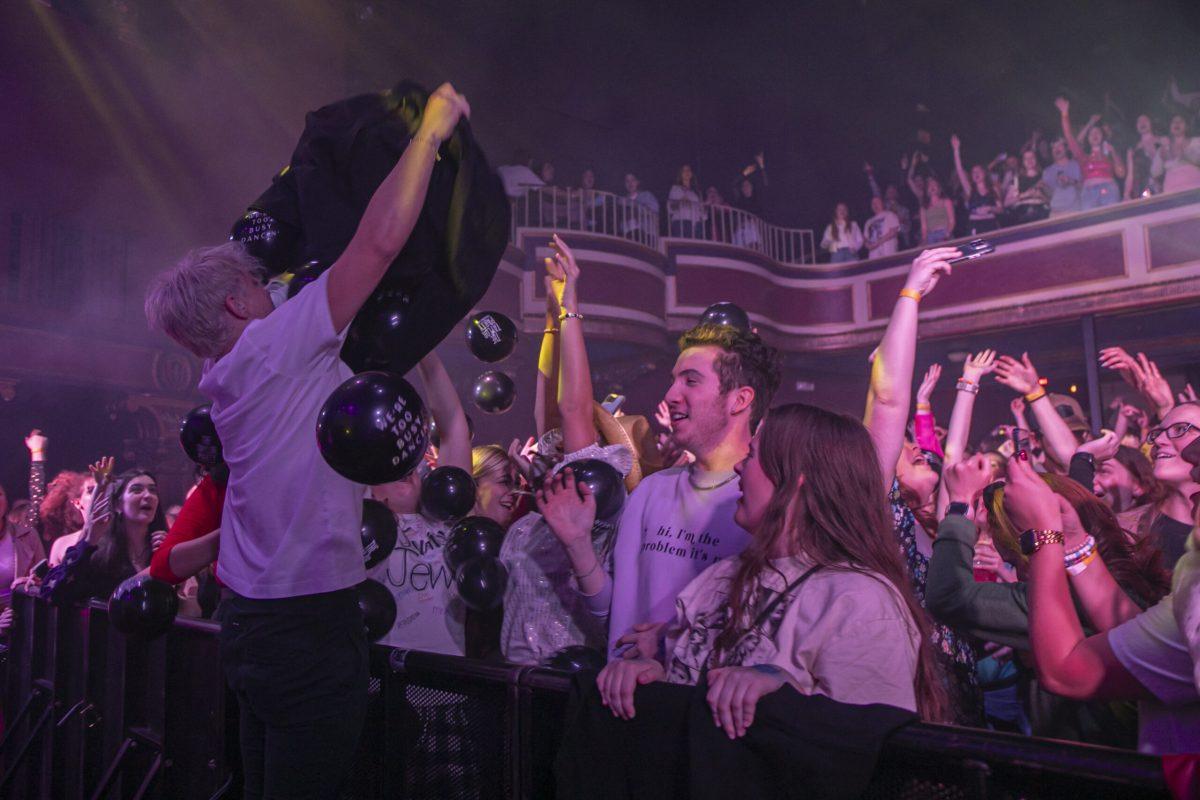 A man, facing a crowd, holds a black bag above his head while balloons fall into the crowd.