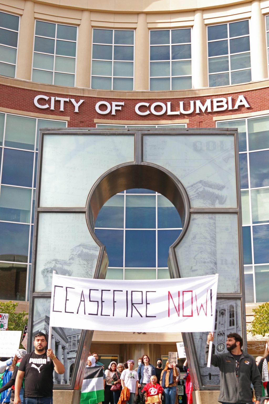 A group of protestors meet outside of a city hall. Two men are holding a banner that says “Ceasefire Now!”
