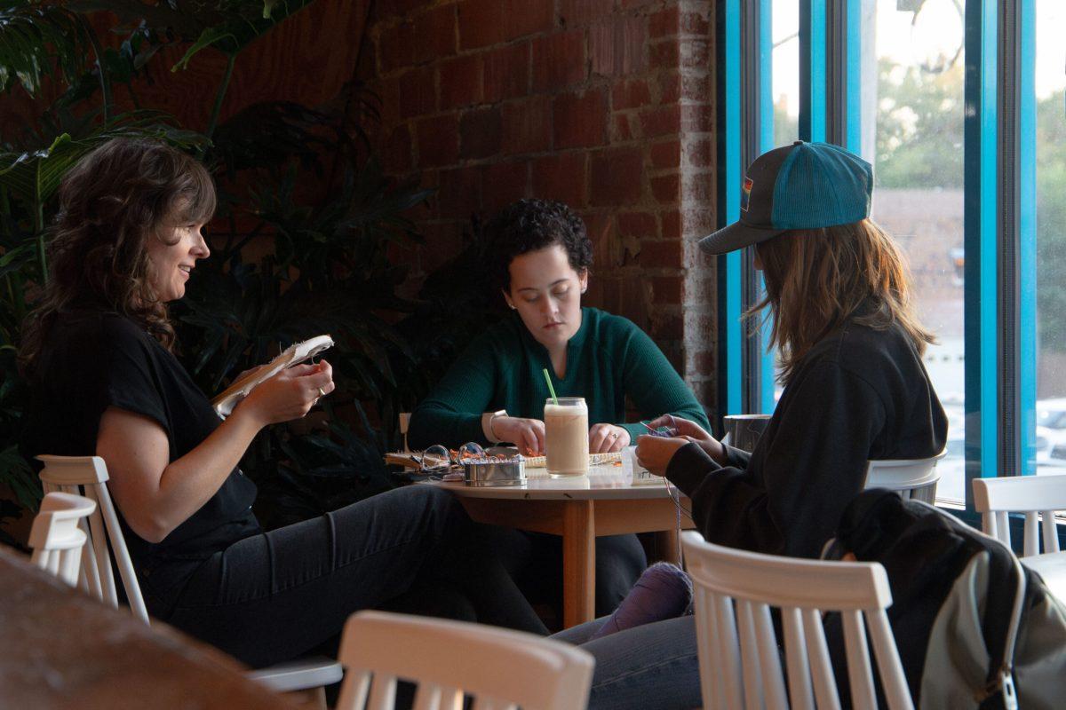Three women sit around a table while working on needle-and-thread crafts.
