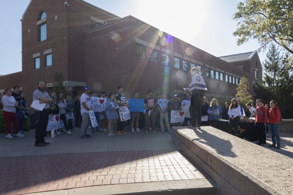 A speaker wearing a yarmulke and the flag of Israel stands in front of a crowd, some holding signs, most saying “We [heart] Israel” with one saying “Israel wants peace! Hamas wants war!”
