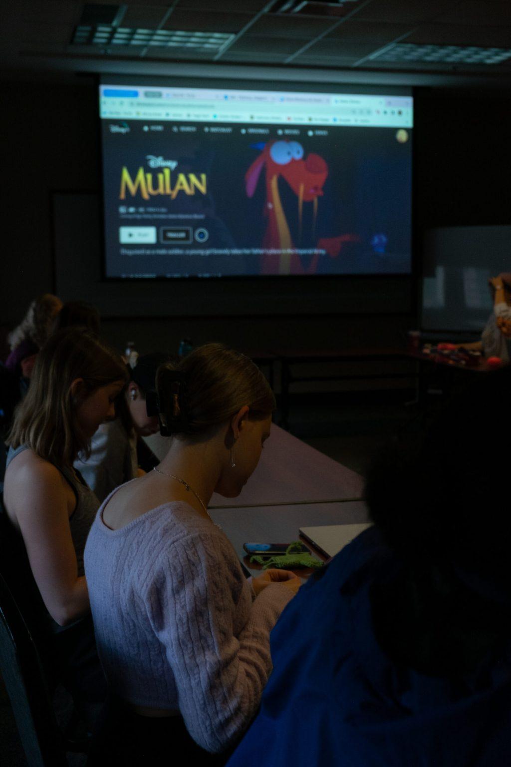 Students sit at tables while the 1998, animated “Mulan” shows on a screen behind them.
