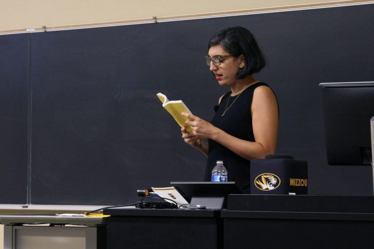 Elif Batuman stands behind a desk reading a book in a lecture hall Tate Hall.
