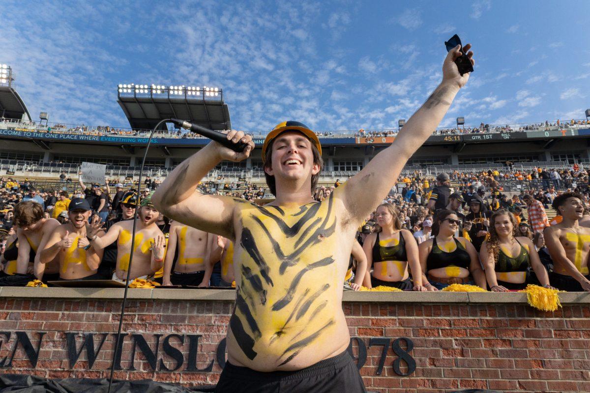 A shirtless man with his chest painted with gold and black stripes holds up a microphone and sunglasses in front of the student section.

