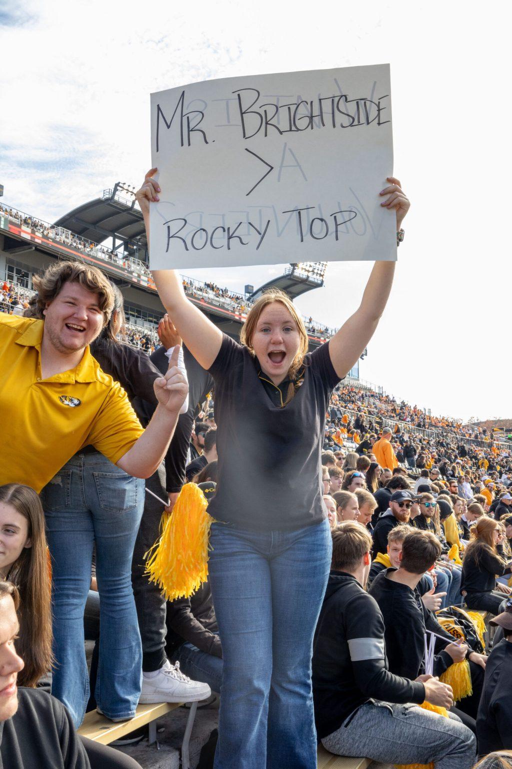 A girl holds up a sign that reads “Mr. Brightside > Rocky Top” as a man holds up a “#1” with his finger.
