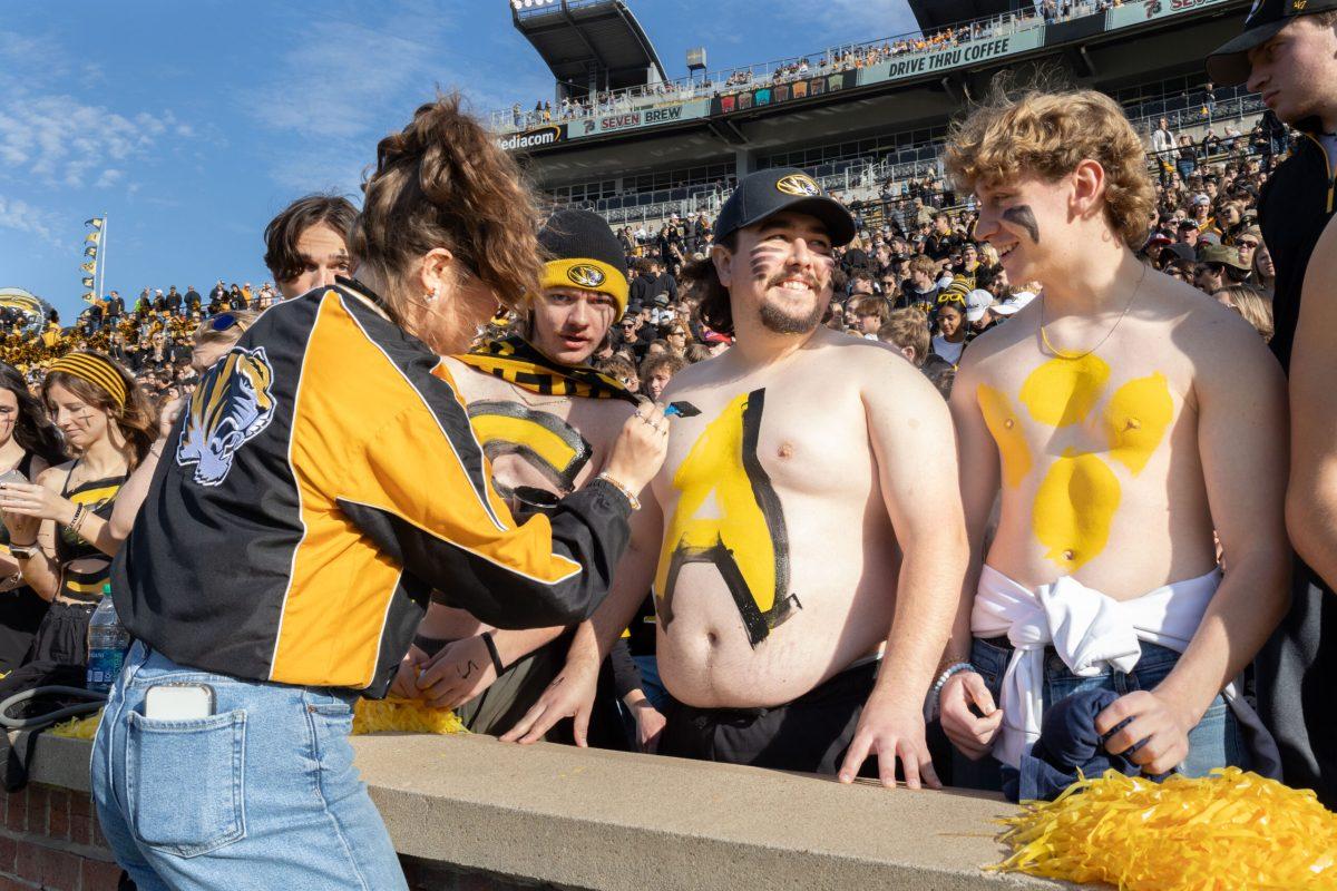 A girl in Missouri merch paints a black and gold “A” onto a man at the front of the student section.
