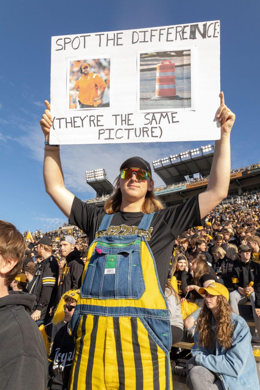 A man wearing gold and black overalls holds up a sign with a picture of the Tennessee coach and a traffic cone with the caption “Spot the difference; they’re the same picture.”
