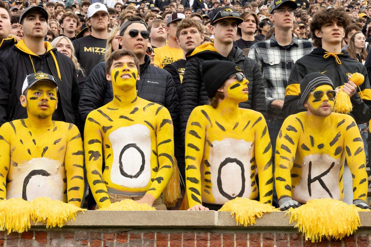 Four men stand at the front of the student section painted gold, with black C-O-O-K painted on their chests.
