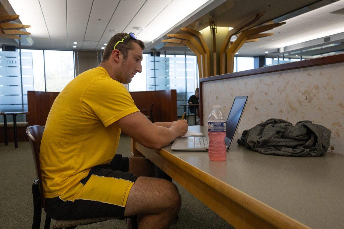 Aidan Dubbert sits in Ellis Library and takes notes in a notebook while looking at his laptop.
