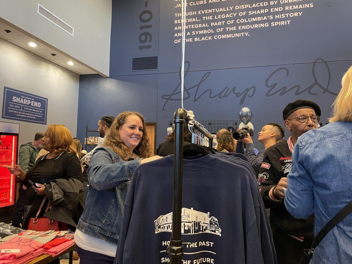 The interior of the Sharp End retail business incubator space. Crowds of people stand between racks of clothing and a high wall is printed with a brief history of the historic Sharp End Black Business District.