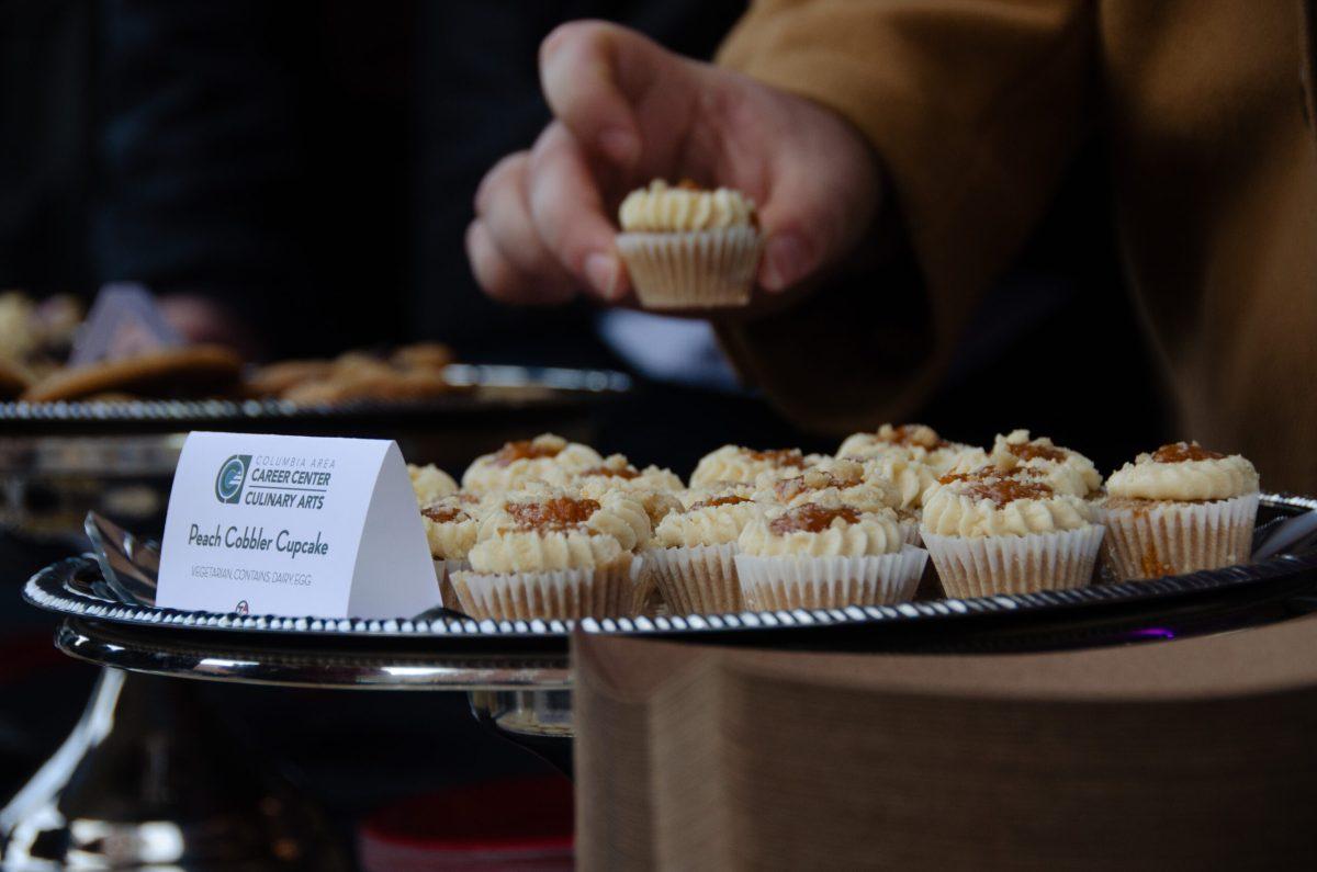 A plate of peach cobbler cupcakes are shown, with a hand reaching to take one.
