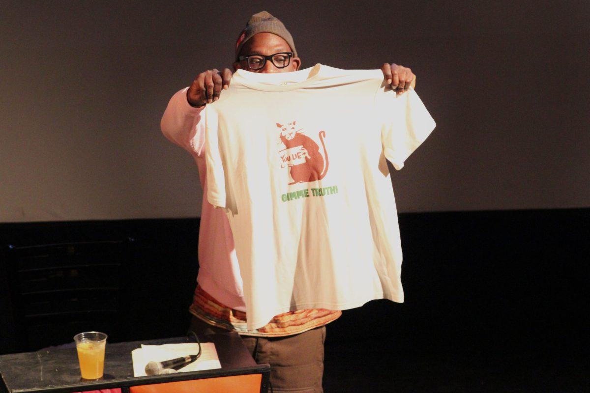 A man holds up a shirt with a rat holding a sign saying “You Lie” and “Gimme Truth” on it.