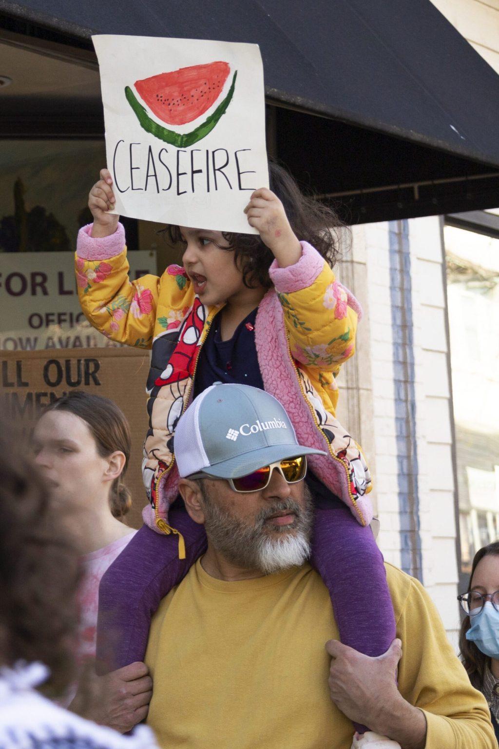 A man holds a young girl up on his shoulders as the young girl holds up a sign with a watermelon on it.
