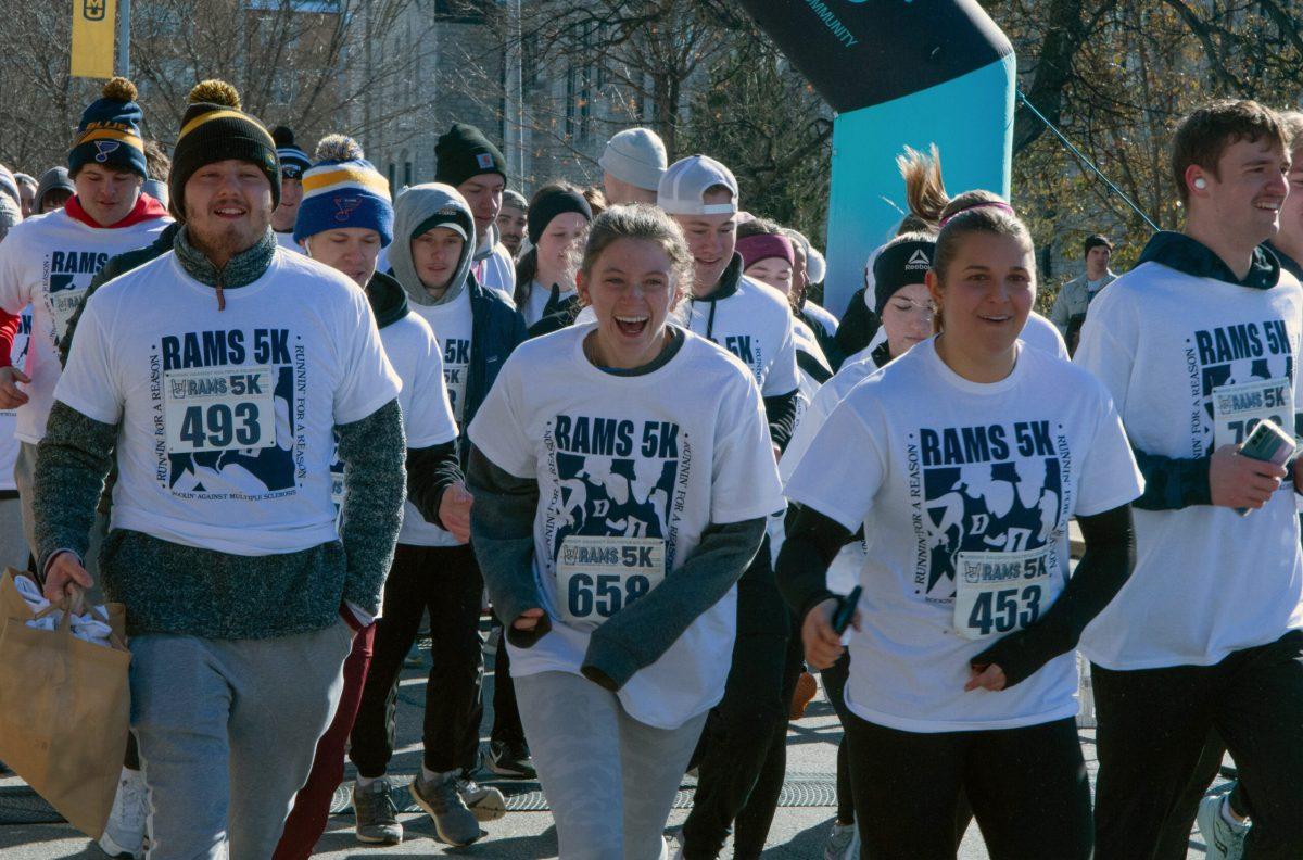 A group of students wearing RAMS 5K shirts and their participant numbers start to run as the 5K begins. The woman in the center of the photo appears to be cold from the wind.