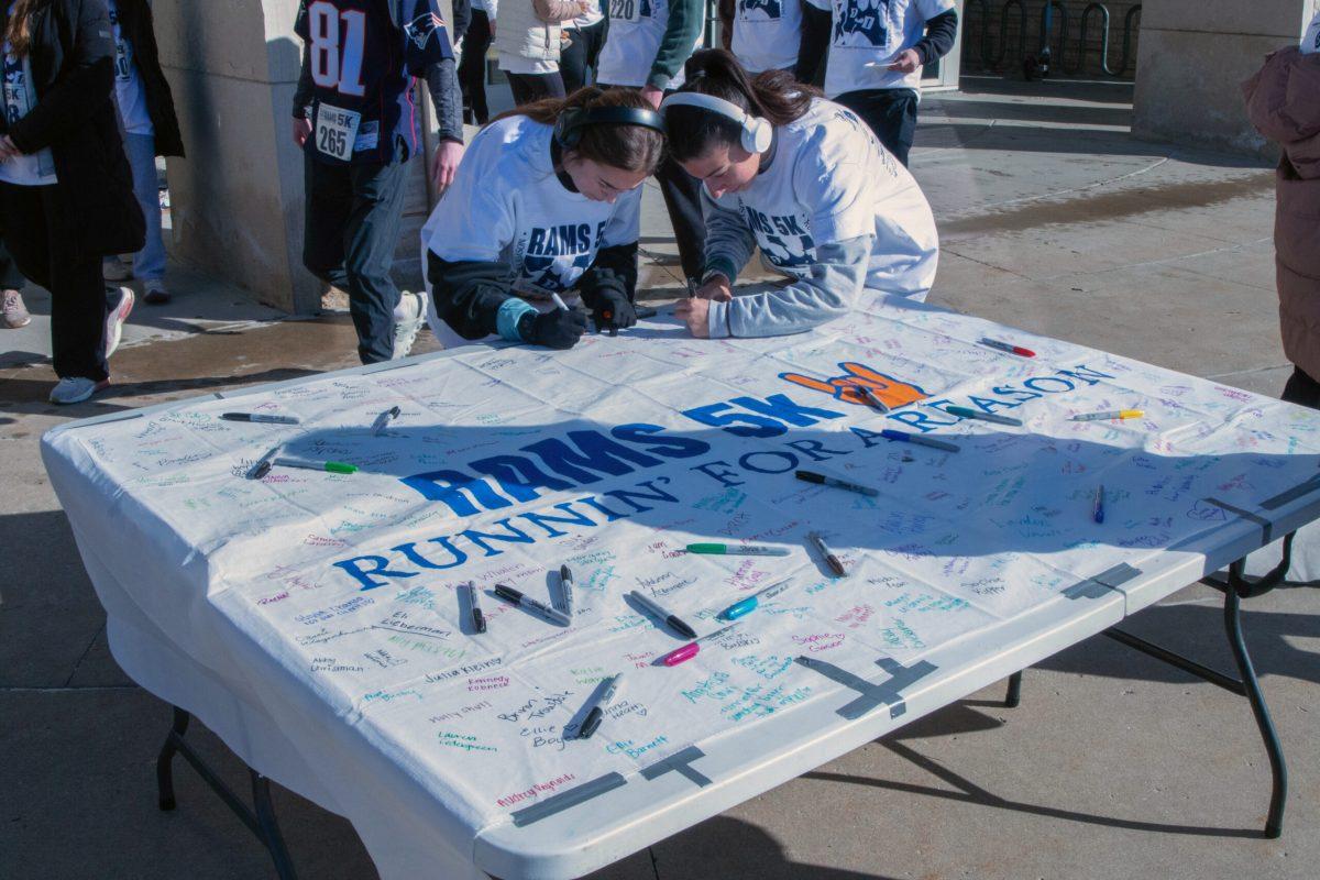 Two women sign their names onto a piece of fabric reading “RAMS 5K Runnin’ For a Reason.” Other signatures can be seen from other students on the fabric.