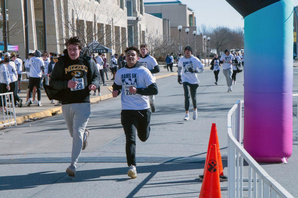 Two males in the foreground running toward the finish line, one in a Mizzou hoodie, and one in a RAMS 5K shirt. There are several runners in the background racing toward the finish.