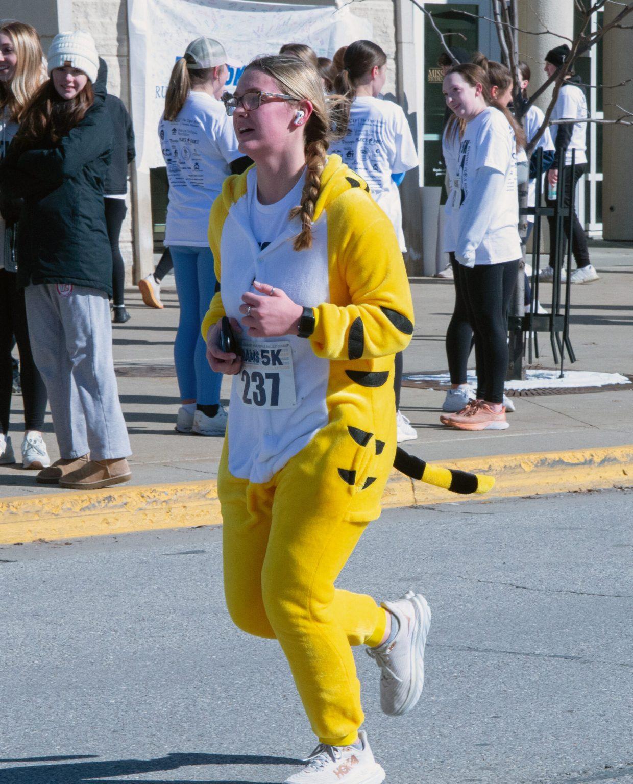 A woman in a yellow tiger onesie with a tail and her participant number attached to her stomach runs in front of the Student Center.