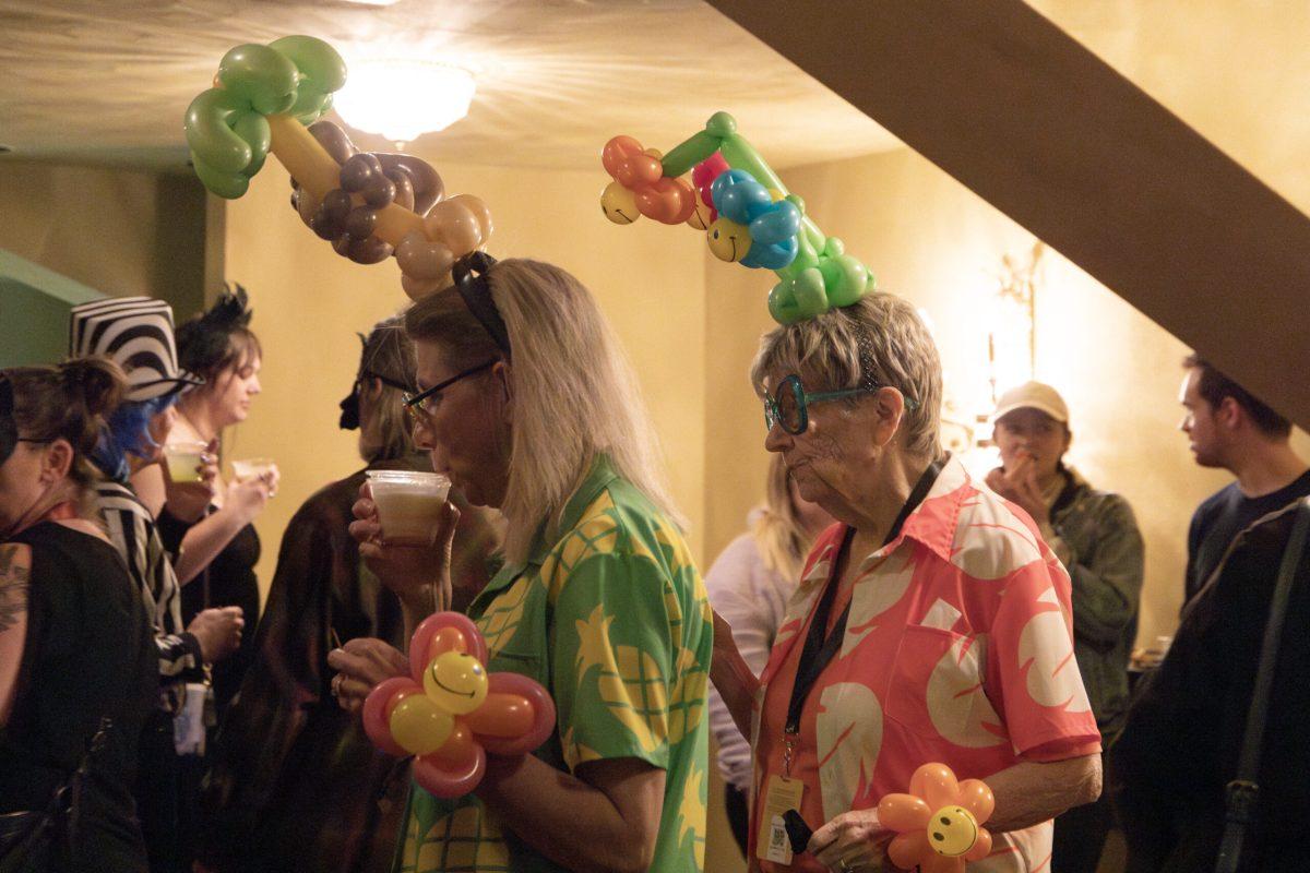 Two women wear balloon hats while holding drinks.
