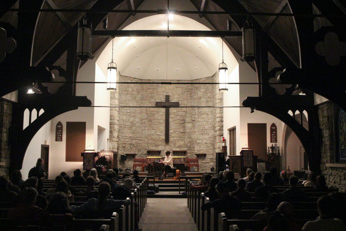 A wide shot of the church with a woman sitting in front of a cross on the stage creating meditative sounds. People are shown in pews on both sides.