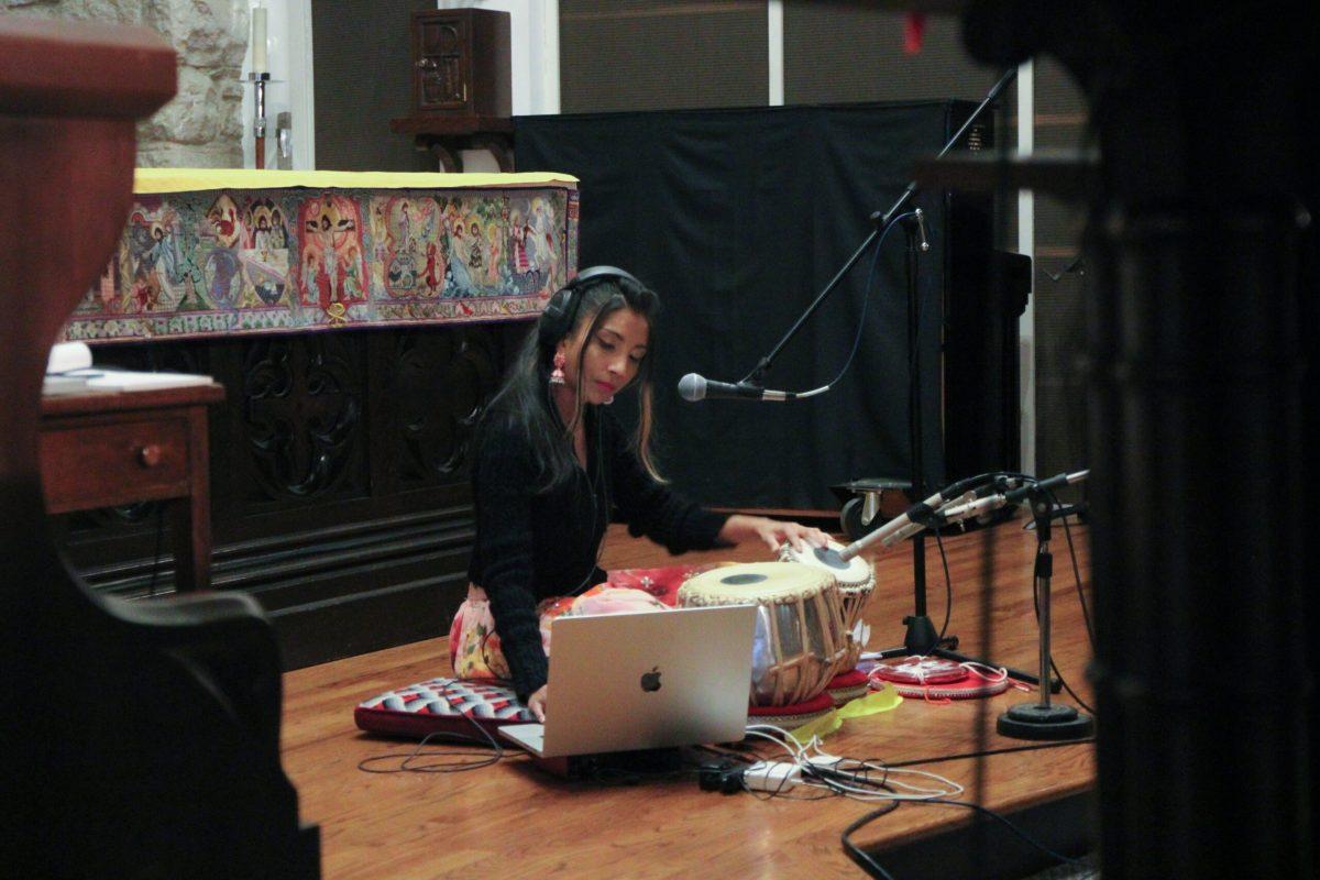 A second woman is shown sitting the stage in front of two tabla drums and is turned towards a MacBook, playing background music.