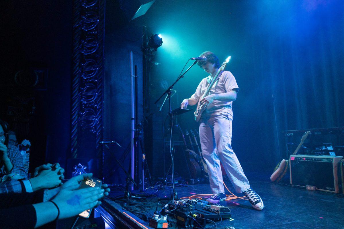 A man with a guitar plays a keyboard on a stage in front of a crowd.
