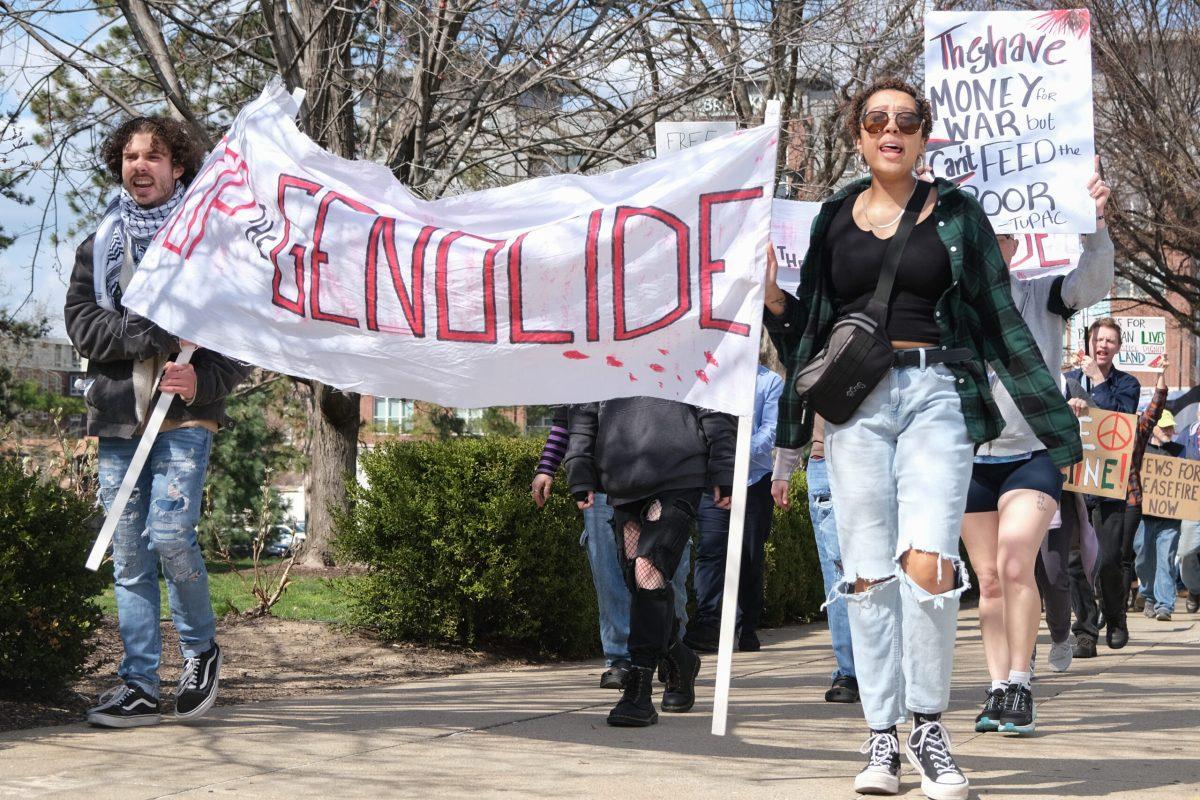 A man and a woman hold a white banner that reads “Stop the Genocide”, and many protesters follow them.