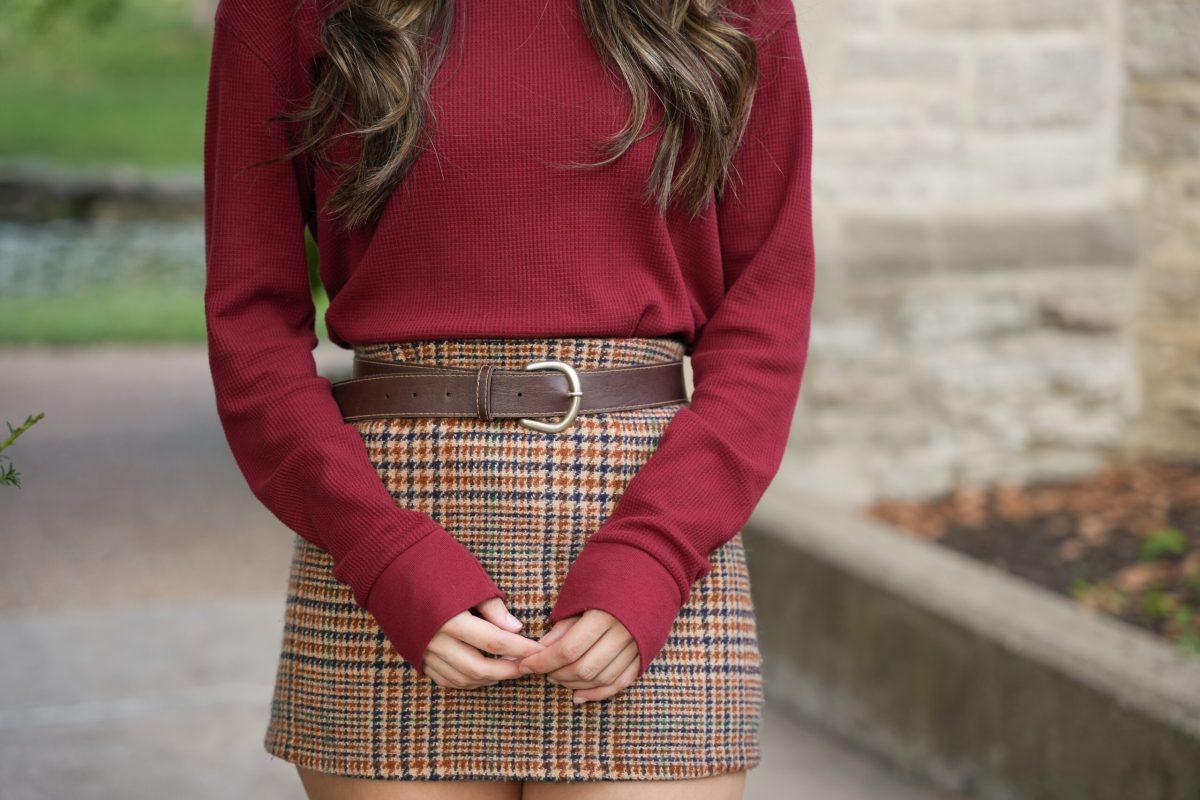 A girl standing dressed in a plaid skirt and burgundy top.