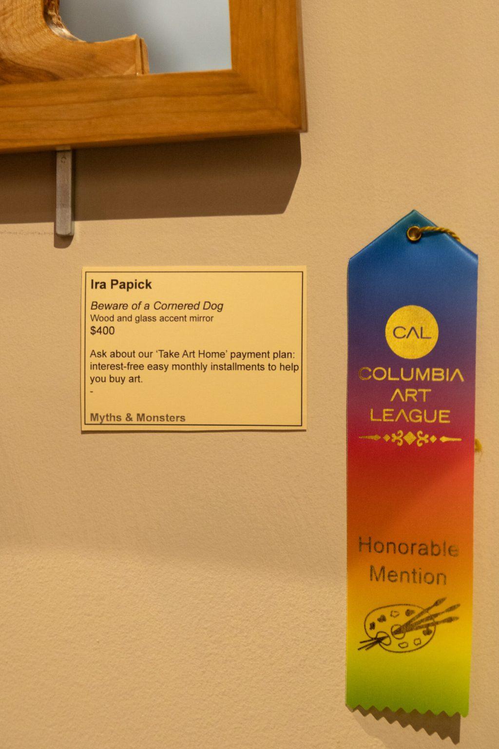 A paper with information about an art piece hanging on a wall next to an honorable mention ribbon.